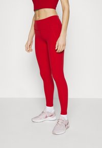 Červené legíny vyrobené z elastického materiálu s vysokým pasem a švy podél stran, spárované se světle růžovými sportovními botami.