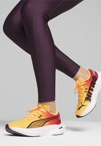 Des chaussures de sport avec un dessus en dégradé jaune et rouge, des accents noirs et une semelle intermédiaire blanche ; portées avec des leggings violets ajustés mettant en valeur les coutures.