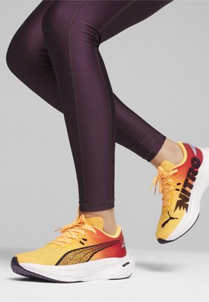 Zapatillas deportivas con una parte superior en degradado amarillo y rojo, acentos negros y una media suela blanca; llevadas con mallas moradas ajustadas que muestran las costuras.