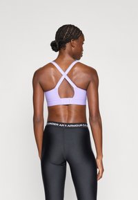 S Sujetador deportivo morado con un diseño de espalda cruzada, que presenta una textura suave y una marca en las correas. Combinado con leggings negros y una cinturilla con logo.