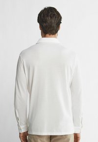 Witte polo met lange mouwen, zachte textuur, knoopsluiting bij de manchetten, subtiele zijslits en een gevouwen kraag. Achteraanzicht getoond.