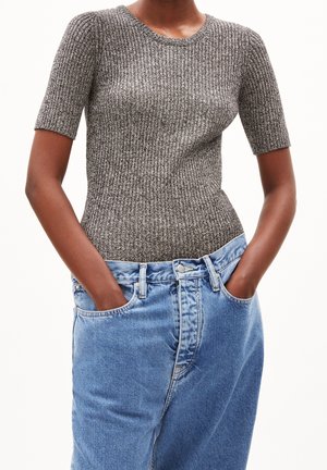 Persoon draagt een nauwsluitend, kort mouwig grijs gebreid shirt, ingestopt in losse, hooggetailleerde blauwe jeans met handen in de zakken.