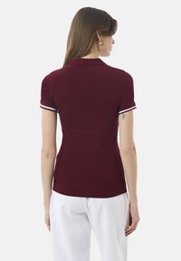 Burgundia polo särk lühikeste varrukatega, valgete ribadega ääred mansettidel ja krael, valmistatud siledast kangast. Tagumine vaade valgete pükstega.