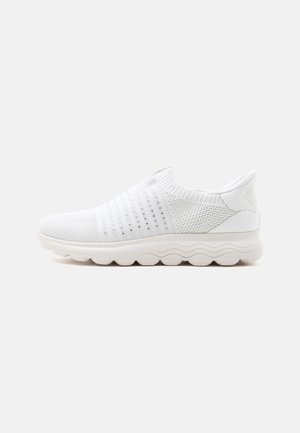 Zapatilla deportiva blanca sin cordones con parte superior de punto, detalles laterales perforados, cuello acolchado y suela ondulada texturizada vista de lado.