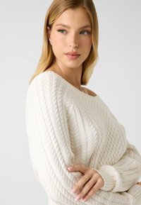 Pull en maille de couleur crème avec un motif torsadé, un large décolleté et des poignets côtelés. Texture douce avec une coupe décontractée.
