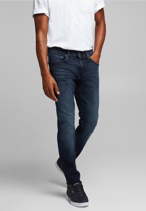 Homme portant un jean slim bleu foncé, une chemise blanche à manches courtes et des baskets noires, debout avec une main dans la poche.