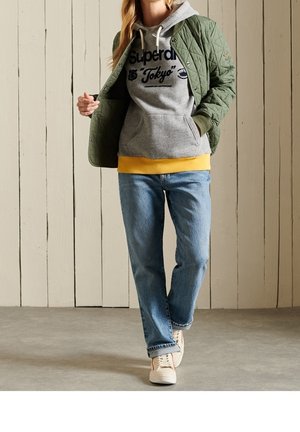 Zelená prošívaná bunda, šedá mikina s kapucí s potiskem „Superdry Tokyo“, žlutý lem, modré džíny a bílé tenisky.