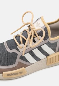 Grå, texturerad tygsneaker med beige accenter, tre vita ränder och beige snören. Har en vadderad sula och en logotyp på sidan.