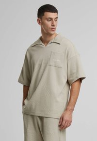 Beige strikket polo shirt med en avslappet passform, korte ermer og en brystlomme med broderte detaljer. Stoffer har et teksturert mønster.