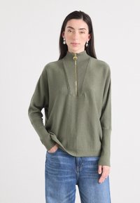 Eine Frau mit langen, dunklen Haaren trägt einen grünen Reißverschlusspullover mit Rippdetail am Kragen, kombiniert mit blauen Jeans und Steckerohrringen, während sie sanft lächelt.