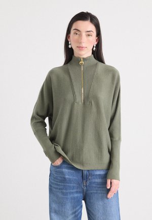 Een vrouw met lang, donker haar draagt een groene ritstrui met ribbels bij de hals, gecombineerd met blauwe jeans en studs in haar oren, terwijl ze zachtjes glimlacht.