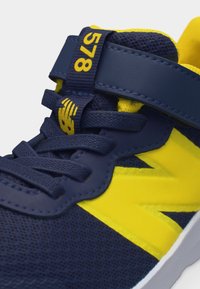 Κοντινή λήψη κορδονιών σε ναυτικό μπλε και κίτρινο χρώμα, λουράκι με το "578" και λεπτομέρειες από υφασμάτινο δίχτυ σε αθλητικό παπούτσι New Balance.