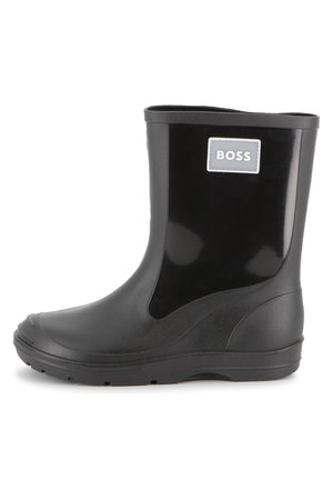 Bottes de cheville en caoutchouc waterproof noires avec une texture lisse et brillante, bout arrondi et un patch logo sur le côté. Elles possèdent une semelle crantée pour une meilleure adhérence.