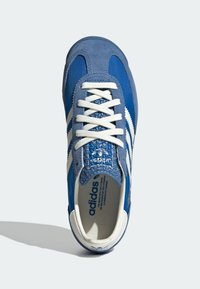 Modrá semišová a látková teniska s bílými pruhy, strukturovanými modrými akcenty a bílými tkaničkami. Má značkovou stélku a detaily na jazyku.