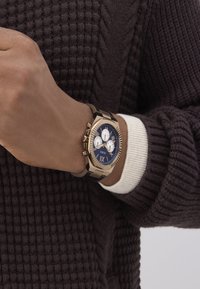 Muñeca con un reloj Guess dorado y negro que presenta una esfera azul y tres subesferas, llevado sobre un puño blanco y un suéter oscuro texturizado.