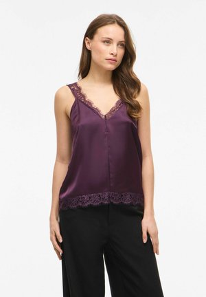 Femme portant un top en satin violet sans manches avec bordure en dentelle au niveau de l'encolure et de l'ourlet, associé à un pantalon noir ample, debout sur un fond blanc.