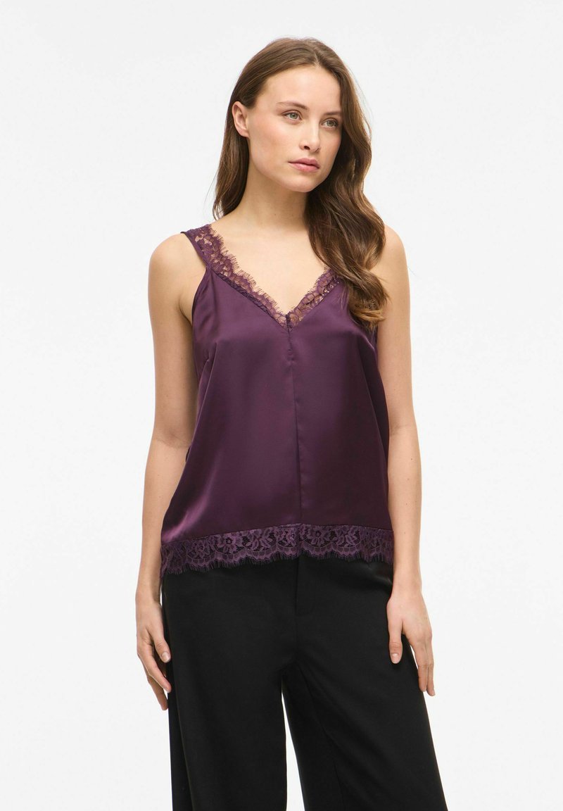 Femme portant un top en satin violet sans manches avec bordure en dentelle au niveau de l'encolure et de l'ourlet, associé à un pantalon noir ample, debout sur un fond blanc.
