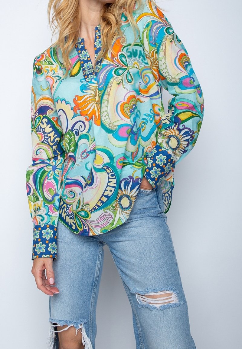 Kvinde iført lyseblå slidte jeans og en iøjnefaldende, langærmet bluse med farverige blomster- og solmønstre.