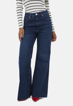 Kobieta ubrana w ciemnoniebieskie, szerokie jeansy z wysokim stanem, biało-czarny paskowany długi rękaw i czerwone, spiczaste buty.