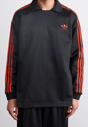 Osoba nosząca czarną koszulkę z długim rękawem z czerwonymi trzema paskami na rękawach oraz małym czerwonym logo Adidas Basketball na piersi.