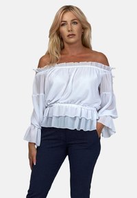 Biała bluzka bez ramiączek z falbanami i długimi rękawami, posiadająca ściągnięty w talii krój i przezroczyste akcenty. Nosić ze spodniach w ciemny, wzorzysty materiał.