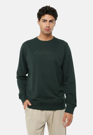 Uomo che indossa una felpa verde scuro con il logo "datch" e pantaloni beige, in piedi con una mano in tasca su sfondo bianco.