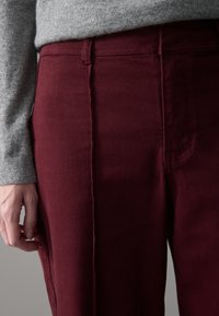 Burgunderfarbene Hose mit glatter Textur, seitlicher Naht und schmaler Passform. Grauer Pullover sichtbar oben, das Material scheint gestrickt zu sein.