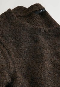 Pruun kootud kampsun tekstuurse materjaliga, millel on kõrge krae ja must silt tekstiga "NEXT" õmmeldud.