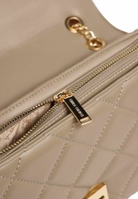 Beige quiltet håndtaske med en guldlås og tilbehør. Indersiden er foret med et mønstret materiale. Lynlåsens trækker har indgraveret mærkenavn.