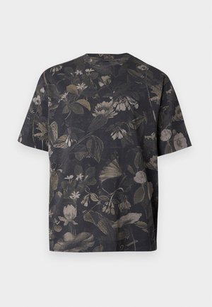 Jeune homme portant un t-shirt à motif floral foncé, un jean bleu clair ample et des baskets blanches, debout devant un fond blanc uni.
