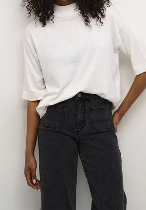 Femme portant un haut à col roulé blanc à manches courtes, ample, rentré dans un jean noir taille haute avec des poches plaquées à l'avant.