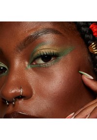Gros plan sur un visage avec un maquillage des yeux vert métallique et or, une peau foncée, et des piercings au septum en argent. Les ongles présentent des accents verts et orange.
