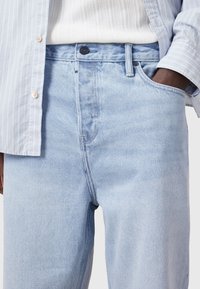 Jeans en denim bleu clair avec une coupe droite, présentant des coutures contrastées et une fermeture à bouton en métal à la taille. Texture lisse, design classique.
