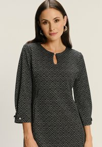 Čierne šaty s diamantovým vzorom, okrúhlym výstrihom s detailom v tvare kľúčovej dierky a rukávmi po tri štvrtiny s gombíkovými akcentmi. Hladká textura.