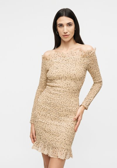 Γυναίκα με μακριά σκούρα μαλλιά που φοράει μια μπεζ φόρεμα με υφή έξω από τον ώμο και διαφανείς βολάν με πουά, ποζάροντας μπροστά σε ένα απλό λευκό φόντο.