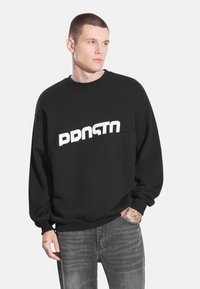 Czarna bluza z białym napisem "PRSTO". Miękka tkanina, luźny krój, ściągacze przy mankietach i u dołu, noszona z szarymi spodniami dżinsowymi.