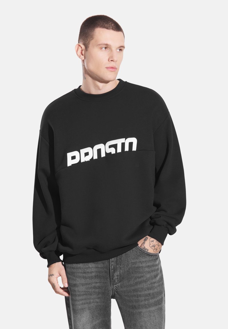 Czarna bluza z białym napisem "PRSTO". Miękka tkanina, luźny krój, ściągacze przy mankietach i u dołu, noszona z szarymi spodniami dżinsowymi.