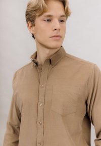 Brun skjorte med knapperekke og krage, enkel brystlomme og teksturert stoff, med frontknapper og en glatt overflate.