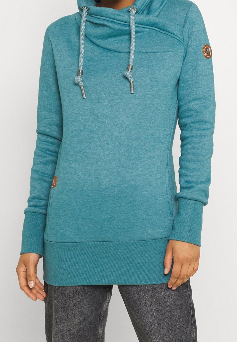 Femme portant un sweat à capuche teal à manches longues avec un col montant et des cordons, associé à un jean gris foncé.