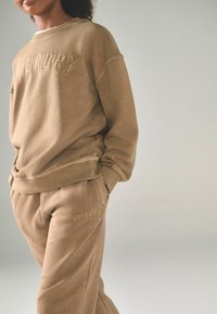 Beige sweatshirt och sweatpants-set med ribbad halsringning och ärmslut. "SUPERDRY" präglad logga på båda plaggen. Mjuk textur.