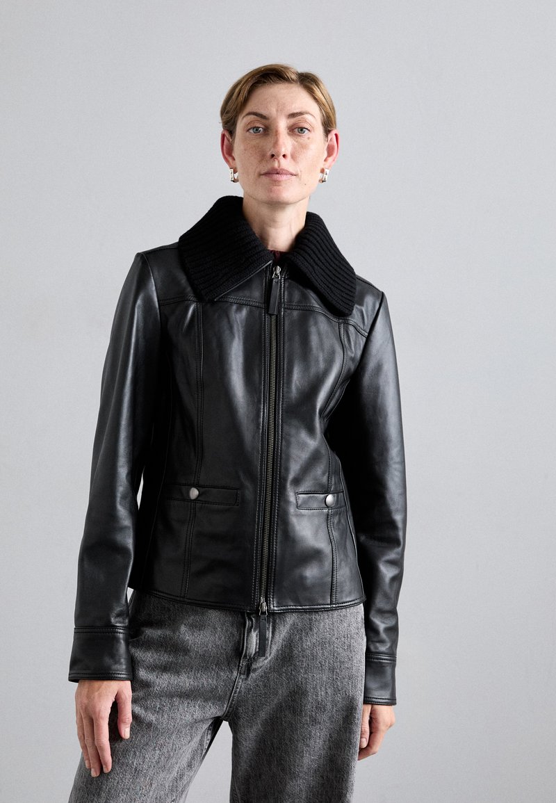 Black leather jacket with a textured black knit collar, front zip, and two side pockets, paired with gray pants. Smooth and structured design.