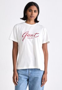 Biała bawełniana koszulka z krótkim rękawem, z okrągłym dekoltem, luźny krój. Na froncie czerwony napis w długopisie "Gant of New Haven". Połączona z jasnoniebieskimi dżinsami.