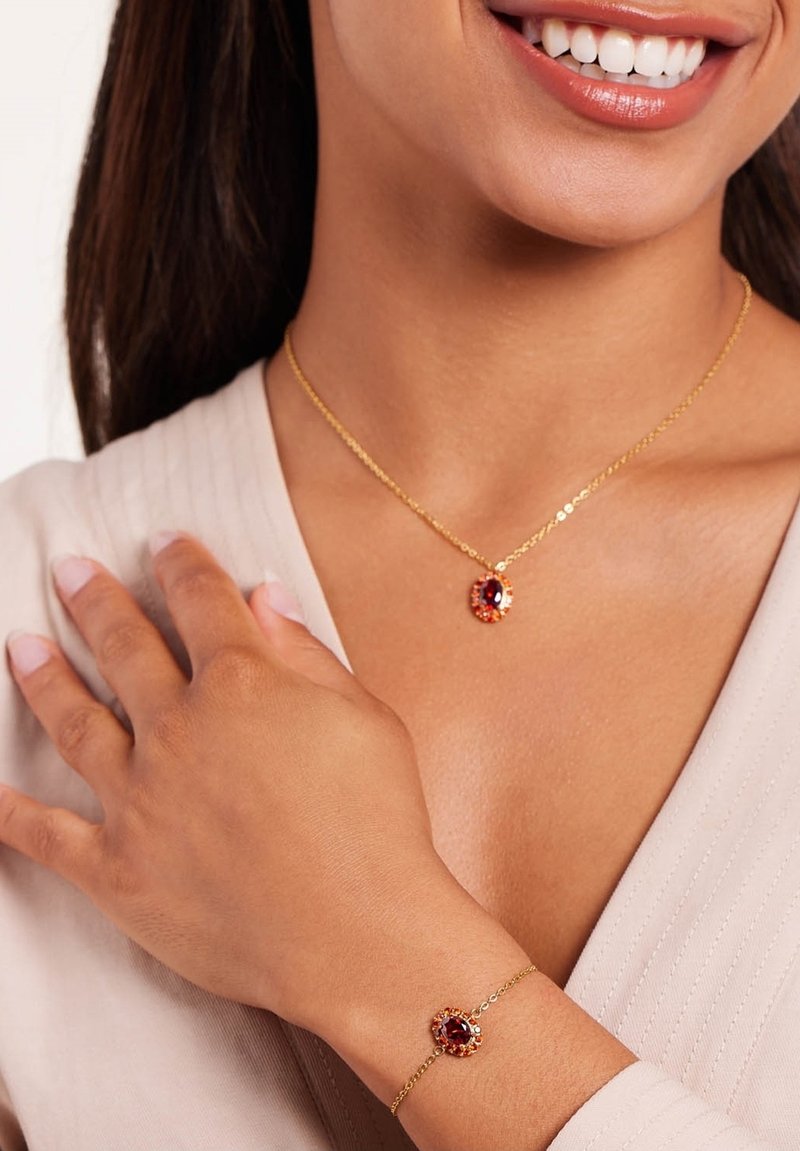 Goudkleurige ketting en armband met een ovale bordeauxrode steen omgeven door kleine heldere stenen, gezet op delicate schakels.