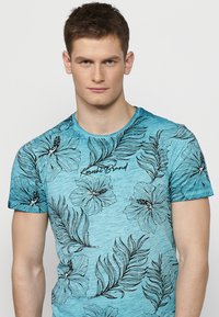 Tirkīza krāsas T-krekls ar īsām piedurknēm, dekorēts ar melniem ziedu un lapu rakstiem. Tam ir brīva piegriezuma forma un apaļa izgriezuma kakla daļa. Zīmola logo izdrukāts uz krūtīm.