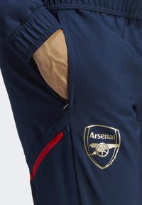 Marineblauwe sportbroek heeft een Arsenal-logo in goud, een helderrode ritszak en een elastische tailleband voor comfort.