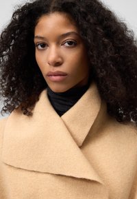 Cappotto di lana beige con un ampio colletto, caratterizzato da una consistenza morbida. Sotto, è visibile un collo alto scuro che completa il colore del cappotto.