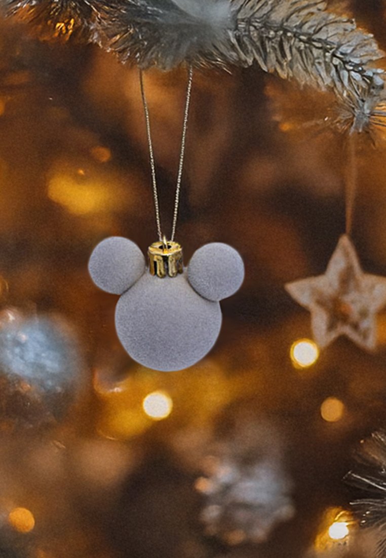 Adorno en forma de Mickey Mouse de terciopelo gris con una tapa dorada para colgar, suspendido frente a un fondo desenfocado de luces festivas en tonos cálidos.