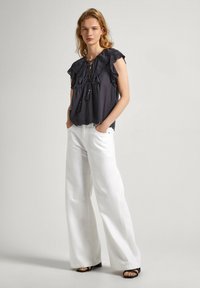 Blusa grigio scuro con maniche a volant, scollo lace-up e frange, abbinata a pantaloni bianchi a gamba larga. Sandali con una sottile fascia visibile.