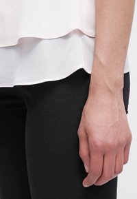 Jasnoróżowa bluzka w warstwach z zaokrąglonym dołem, noszona na czarnych spodniach. Widoczna ręka spoczywająca na boku, ukazująca teksturę skóry i detale.