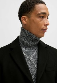 Blazer in misto lana nera su un maglione lavorato a maglia grigio a collo alto con un motivo testurizzato. Primo piano, in evidenza i dettagli e il contrasto tra i colori.
